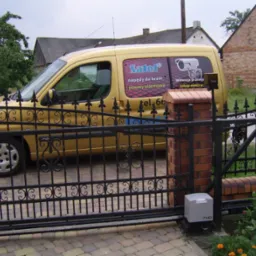 Złoty samochód dostawczy z reklamą firmy instalującej napędy do bram i systemy alarmowe, zaparkowany za czarną, kutą bramą z automatycznym napędem FAAC.