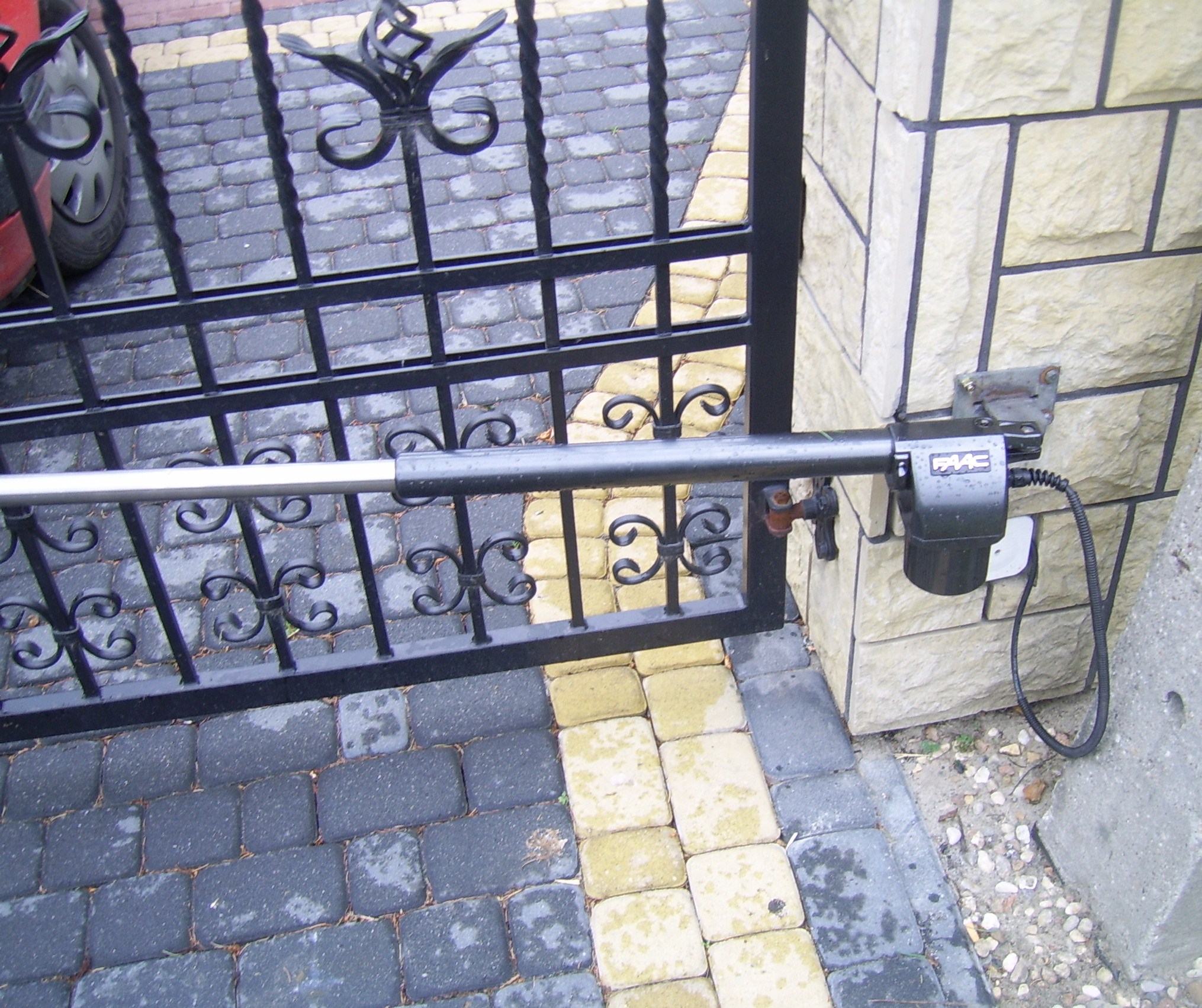 Automatyka bramy: siłownik FAAC zamontowany przy ozdobnym, kutym ogrodzeniu posesji z dwukolorowej kostki brukowej