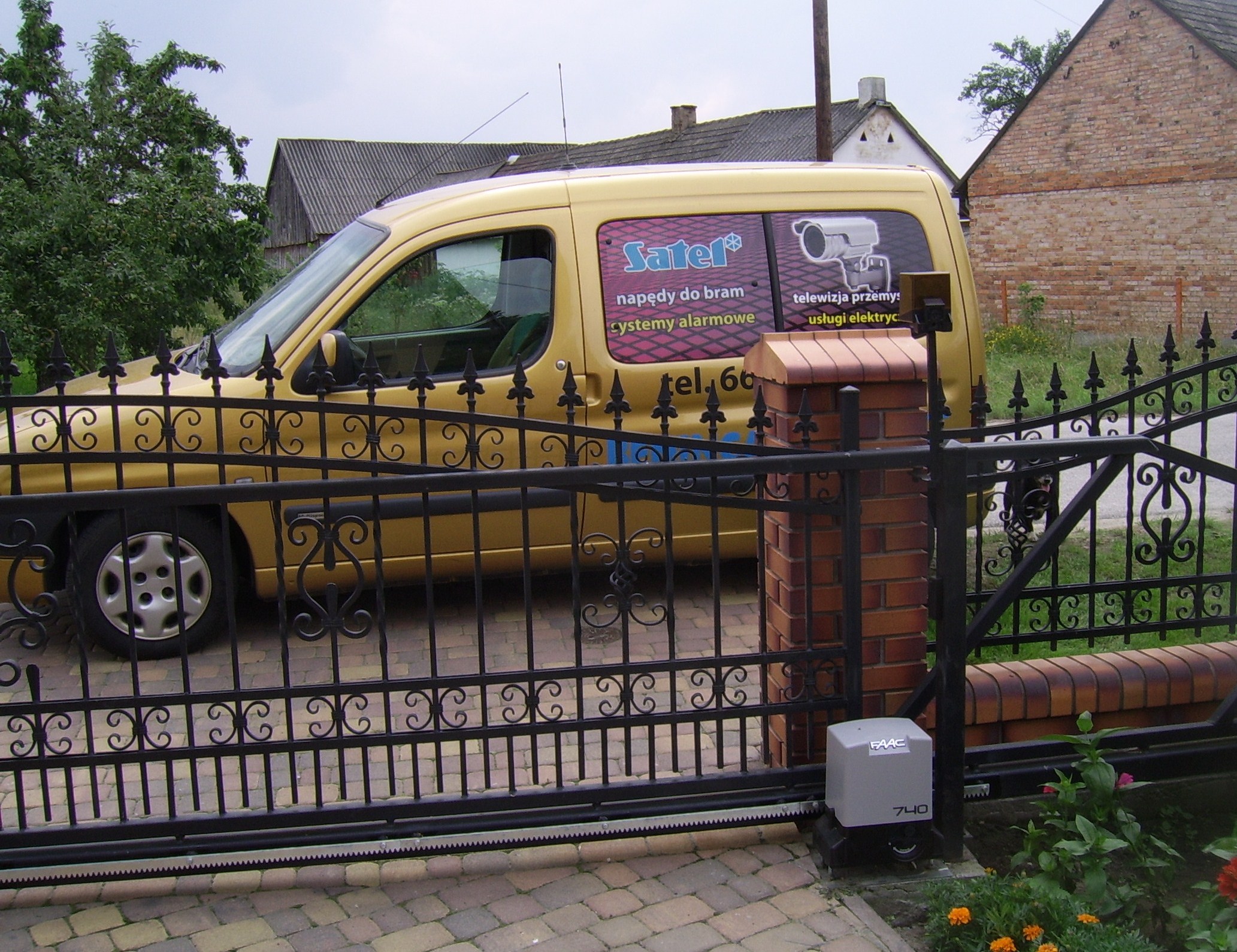 Złoty samochód dostawczy z reklamą firmy instalującej napędy do bram i systemy alarmowe, zaparkowany za czarną, kutą bramą z automatycznym napędem FAAC.