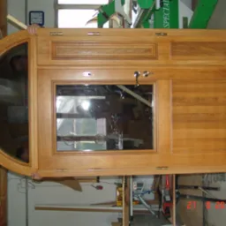 Drewniane drzwi z szybami o nietypowym, półokrągłym kształcie, częściowo trzymane przez dwie osoby, widoczne elementy warsztatu w tle.