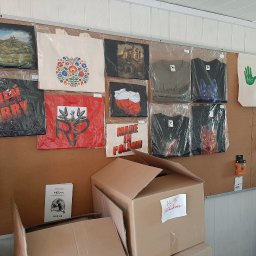 Hifilon Drukarnia DTF - Korkowa tablica z ekspozycją różnorodnych, pakowanych koszulek i toreb bawełnianych z nadrukami: zamki, wzory ludowe, patriotyczne i zwierzęce grafiki.