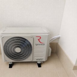Mikołaj Nowak - Zewnętrzna jednostka systemu chłodzącego Rotenso Inverter, zamontowana na beżowych płytkach przy jasnej, teksturowanej ścianie, z widocznymi przewodami.