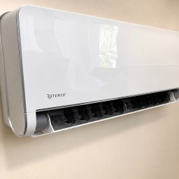 Mikołaj Nowak - Biała, nowoczesna jednostka wewnętrzna klimatyzatora ściennego Rotenso, zamontowana na jasnobeżowej ścianie. Widoczne czarne lamele wylotu powietrza i odbicie okna.