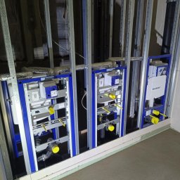 INSTALATORSTWO HYDRAULICZNE SEBASTIAN KRAWCZYK - Trzy niebieskie stelaże podtynkowe Geberit do WC, zamontowane w konstrukcji z metalowych profili, z widocznymi rurami i izolacją, przed zabudową ścian w łazience.