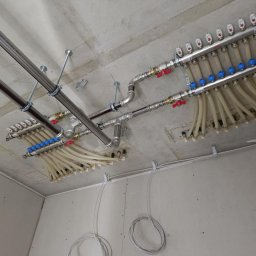 INSTALATORSTWO HYDRAULICZNE SEBASTIAN KRAWCZYK - Dwa chromowane rozdzielacze instalacji grzewczej z licznymi elastycznymi rurami i zaworami, zamontowane na betonowym suficie. Widoczne główne rury zasilające i okablowanie na ścianach z płyt GK.