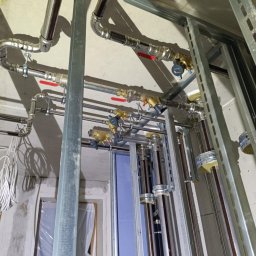 INSTALATORSTWO HYDRAULICZNE SEBASTIAN KRAWCZYK - Lśniące rury instalacji grzewczej i wodnej z mosiężnymi zaworami oraz licznikami, zamocowane na metalowych profilach konstrukcyjnych w trakcie prac.