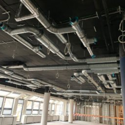 INSTALATORSTWO HYDRAULICZNE SEBASTIAN KRAWCZYK - Srebrne kanały wentylacyjne i okablowanie elektryczne na ciemnym suficie w dużym, nieukończonym pomieszczeniu komercyjnym z szerokimi oknami i białymi kolumnami.