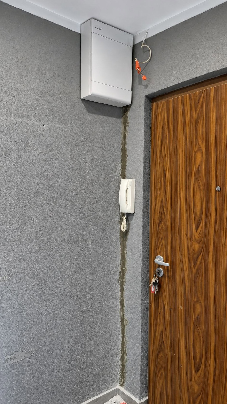 Biała skrzynka elektryczna Hager i domofon na szarej, teksturowanej ścianie, obok drewnianych drzwi z kluczami. Widoczne przewody z pomarańczowymi złączkami i świeże wypełnienie szczeliny w narożniku.