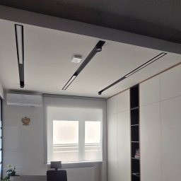 Budolid - Nowoczesne wnętrze z białymi ścianami i sufitem, wyposażone w czarne, liniowe oświetlenie szynowe LED. Widoczne okno z roletą, klimatyzator i elegancka zabudowa meblowa.