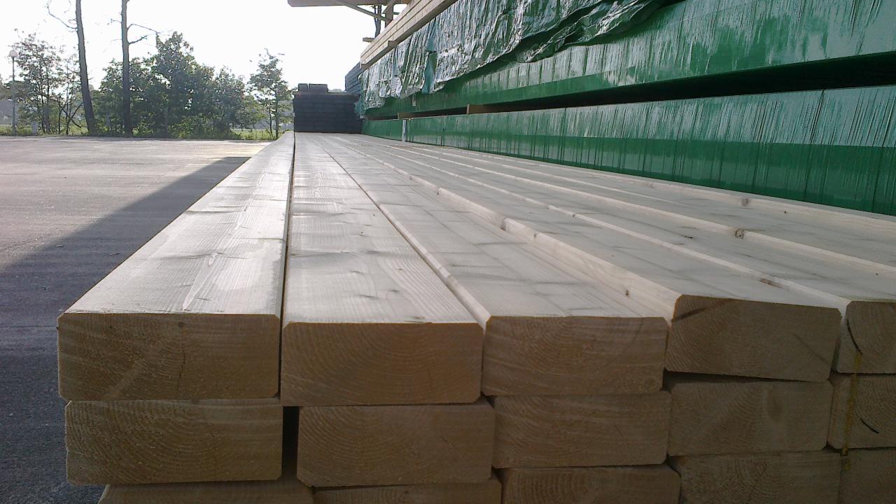 Stos tarcicy budowlanej ułożony na zewnątrz, z zieloną plandeką w tle. Widoczne słoje drewna i krawędzie belek. Jasne, naturalne światło.