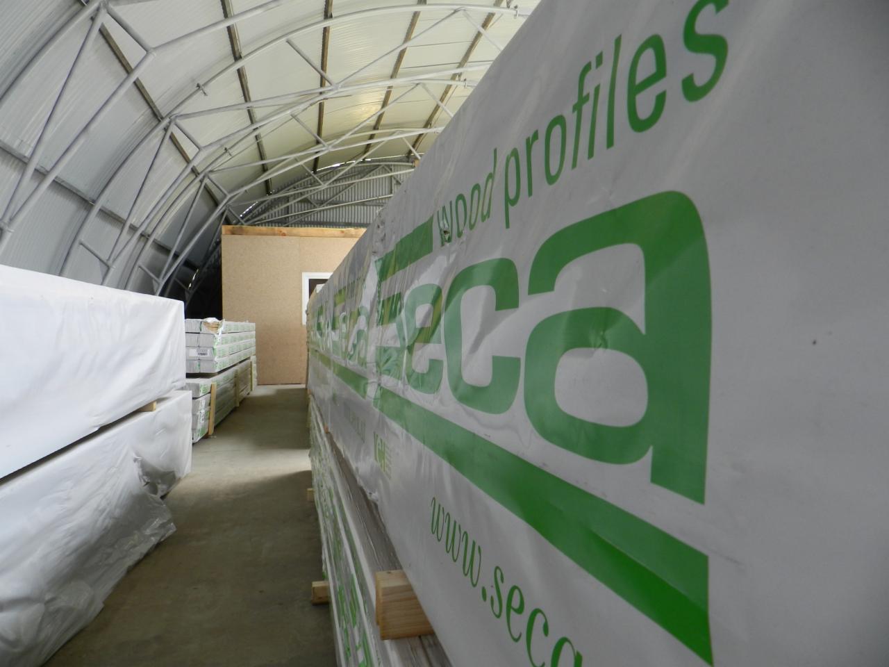 Wnętrze magazynu z belami tarcicy budowlanej SECA, owiniętej w białą folię. Widoczne profile i logo w zieleni. Metalowa konstrukcja hali w tle.
