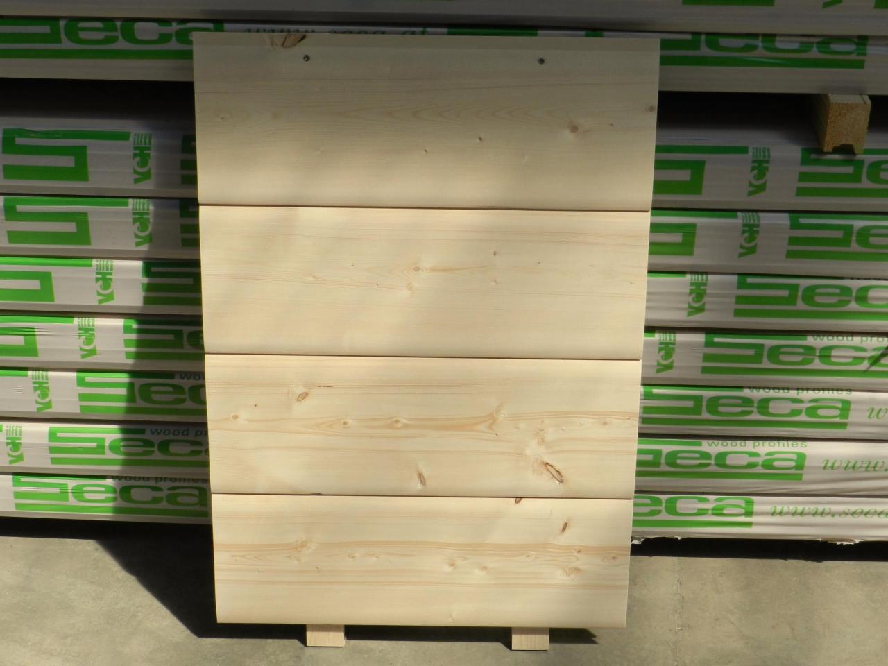 Deska elewacyjna z jasnego drewna sosnowego na tle opakowań z logo producenta, eksponowana w naturalnym świetle. Widoczne słoje i niewielkie sęki.