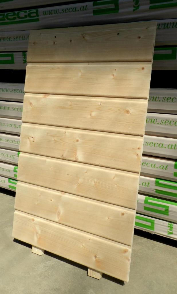 Jasna deska elewacyjna z widocznym rysunkiem drewna, oparta o podłoże na małych klockach, na tle pakietów desek z zielonym nadrukiem.