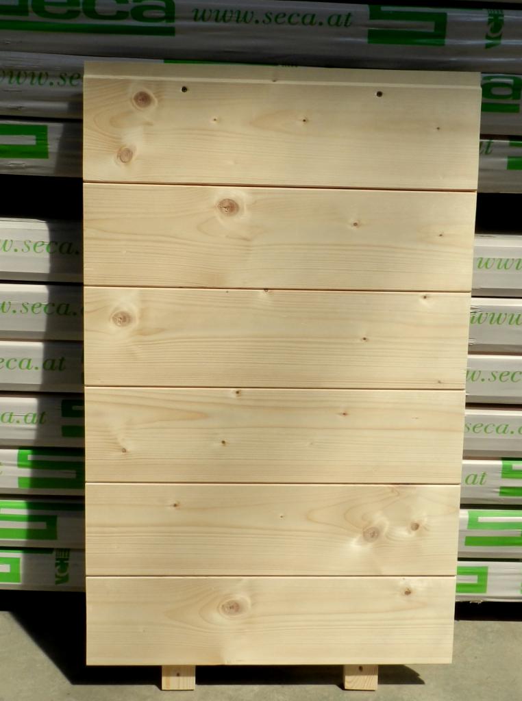 Jasna, sosnowa tarcica z Wilkowic, ułożona poziomo. Widoczne słoje i niewielkie sęki. Całość wsparta na małych, drewnianych nóżkach. W tle opakowania tarcicy.