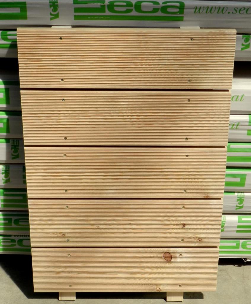 Drewniana konstrukcja z desek, jasne drewno z widocznymi śrubami, na tle opakowań z zielonym nadrukiem, prosta forma, naturalny materiał.