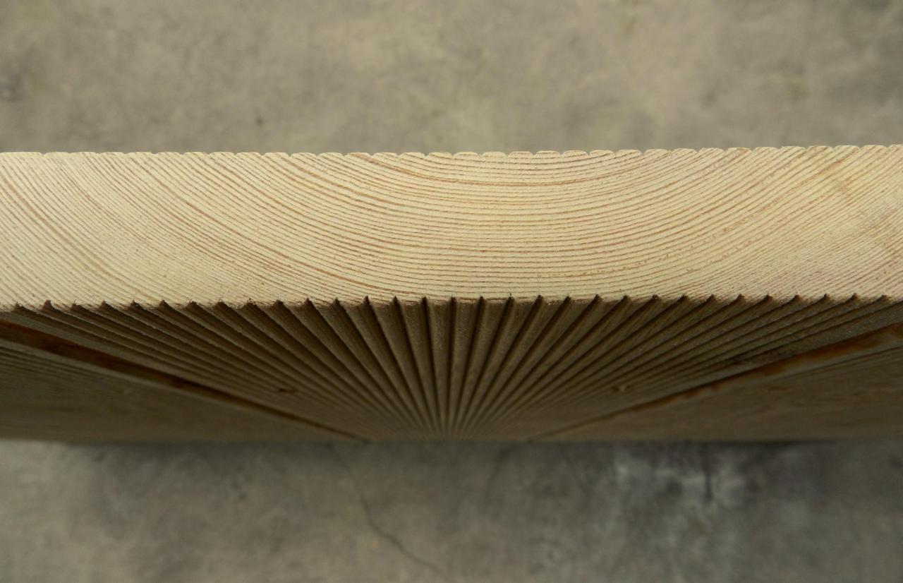 Detal drewna z wyfrezowanym wzorem, ukazującym słoje. Widoczne precyzyjne nacięcia i jasny kolor drewna. Surowe wykończenie, bliski kadr.