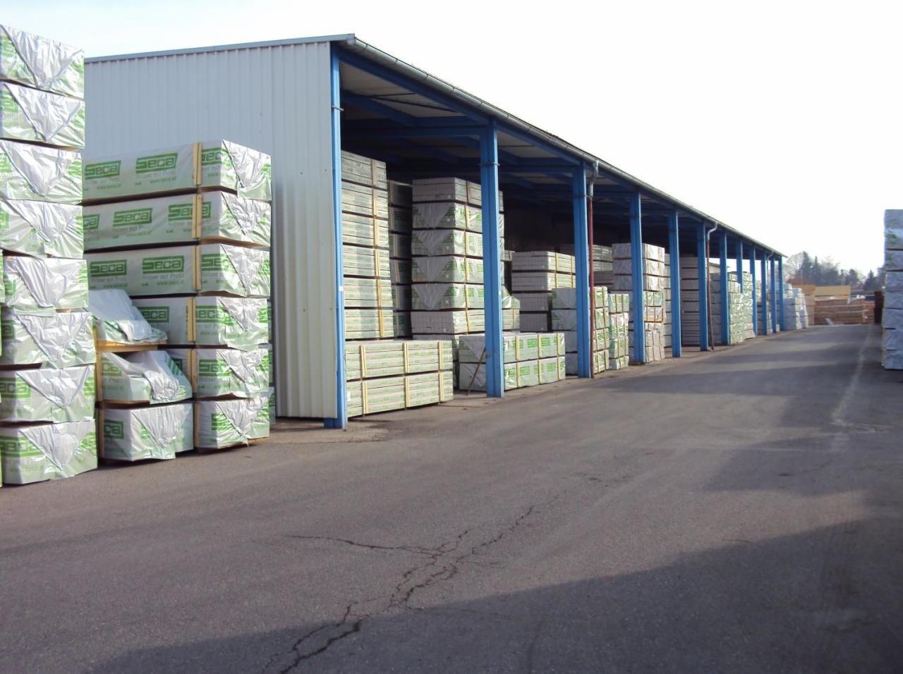 Składowana tarcica budowlana w magazynie z niebieskimi słupami. Drewno ułożone w rzędach, zabezpieczone folią z zielonymi napisami. Widok z perspektywy drogi dojazdowej.