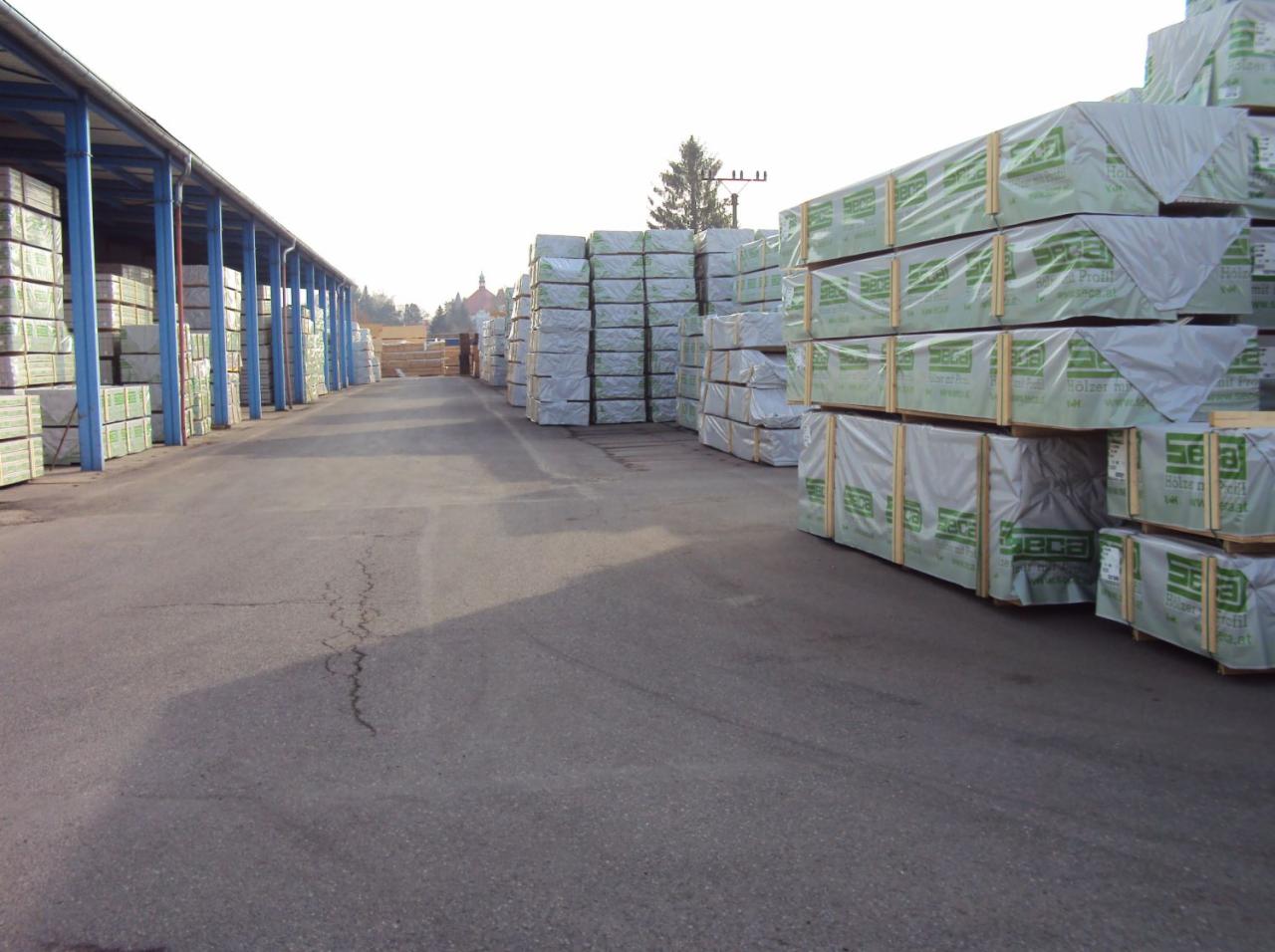 Składowisko tarcicy: rzędy spakowanych pakietów drewna na paletach, częściowo pod zadaszeniem, z widocznym budynkiem w oddali i drzewem na horyzoncie.