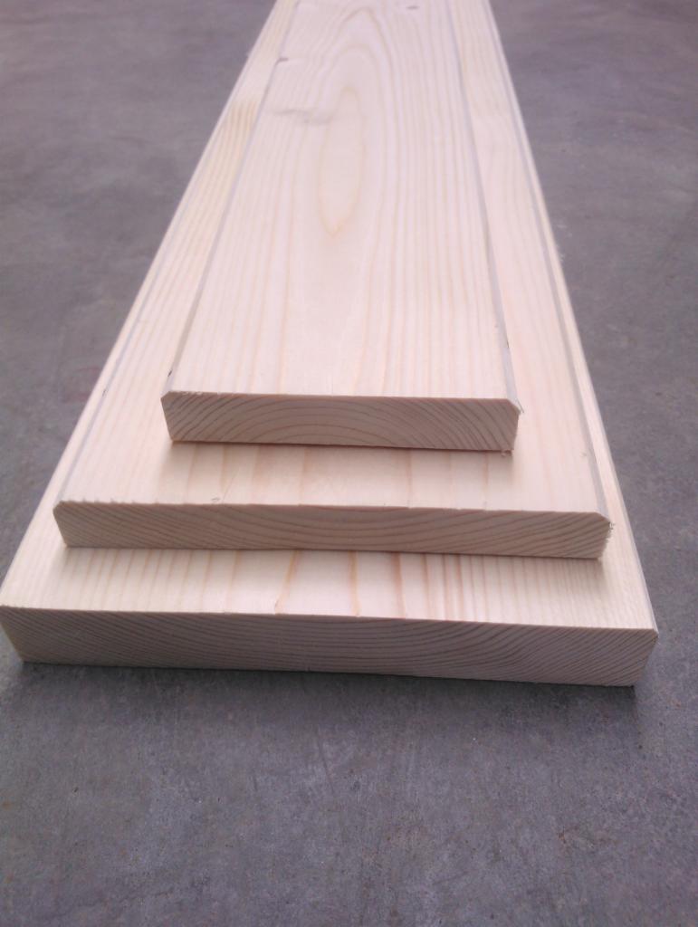 Surowe, jasne deski ułożone warstwowo na szarym tle. Widoczne słoje drewna i precyzyjne cięcia. Deska na wierzchu posiada delikatne frezowanie. Widok z perspektywy.