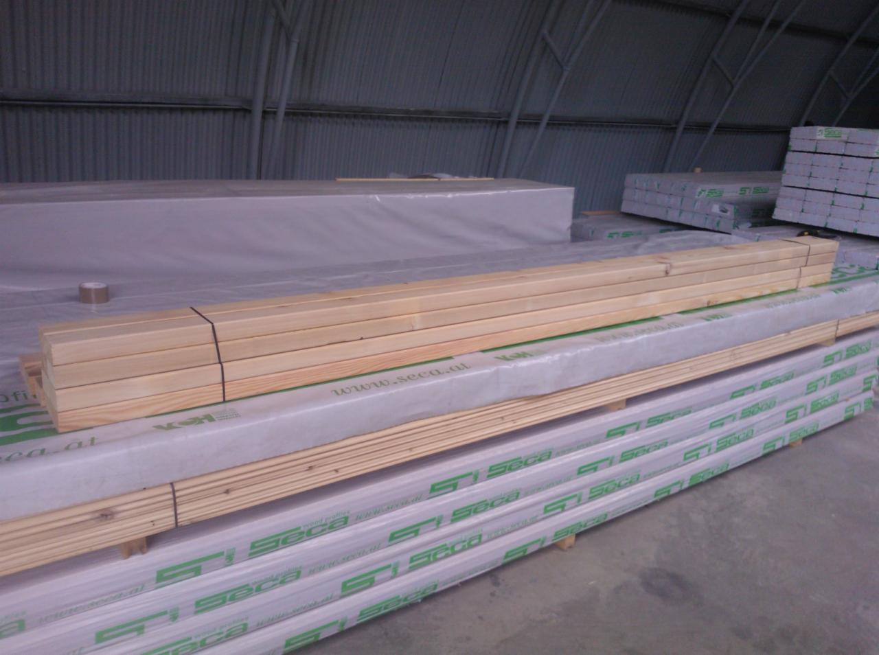 Drewniane belki spięte czarnym sznurkiem leżące na palecie z logo 'Seca' w magazynie z blachy falistej. W tle inne palety z belkami.