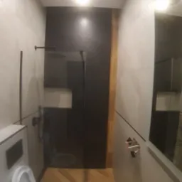 Nowoczesna łazienka z czarną kabiną prysznicową typu walk-in, białą ceramiką i podłogą z parkietu ułożonego w jodełkę, oświetlona jasnym światłem.