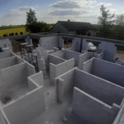 Wewnętrzne ściany domu w trakcie budowy wzniesione z białych bloczków, widoczne rusztowania i samochód w tle, żółte pole rzepaku i dom w oddali.