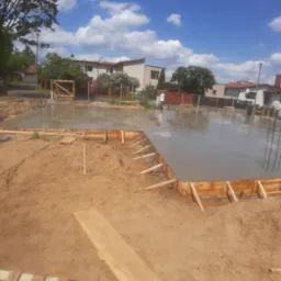 Wylany fundament z drewnianym szalunkiem w kształcie prostokąta na tle domów jednorodzinnych i błękitnego nieba.