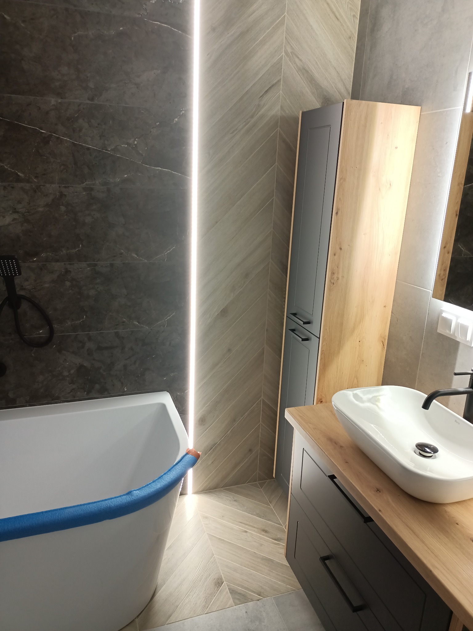 Nowoczesna łazienka z wanną zabezpieczoną niebieską pianką, umywalką nablatową na drewnianym blacie i szafką w odcieniach szarości i drewna, podświetlana listwa LED na ścianie.
