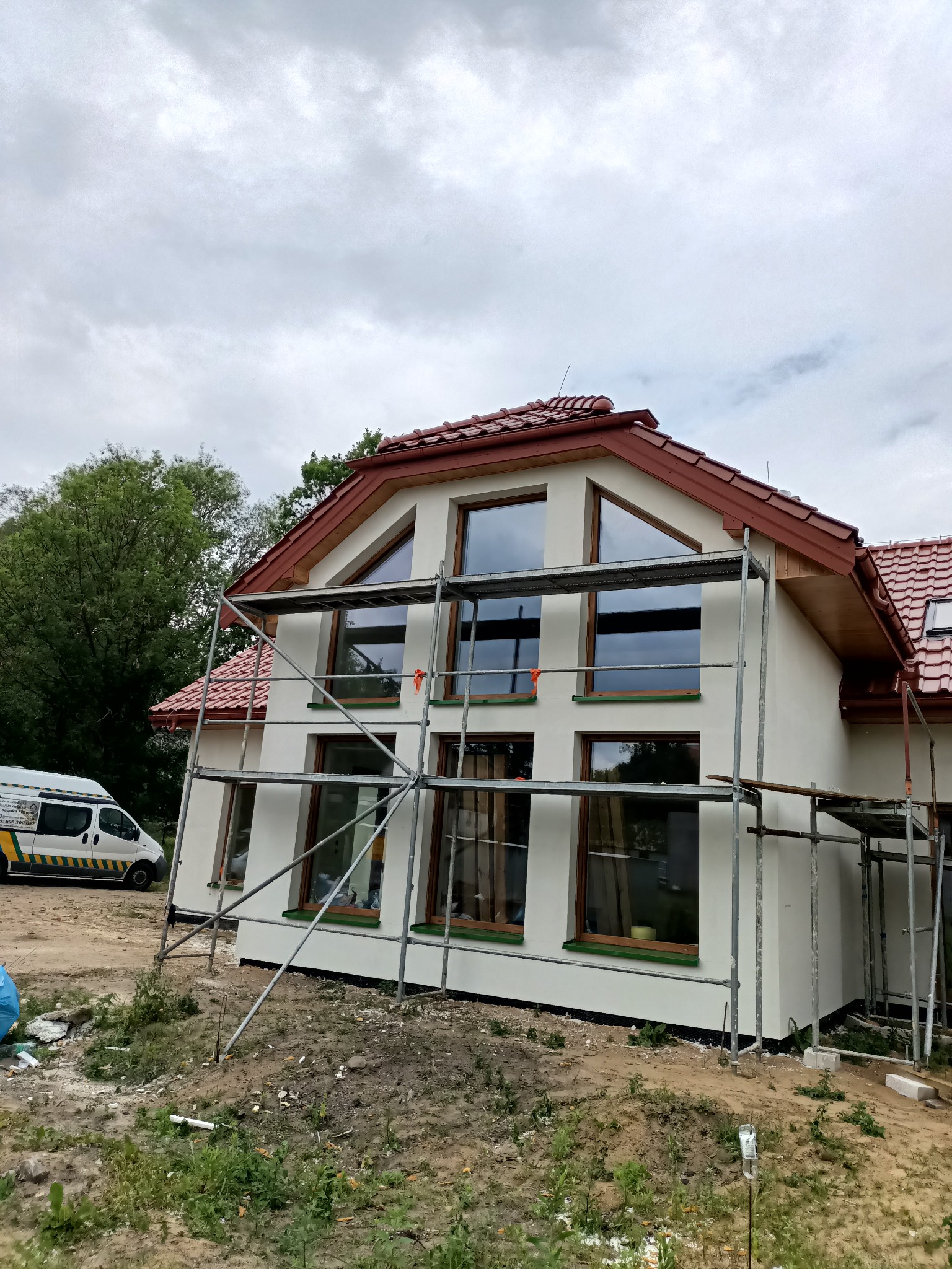 Dom jednorodzinny z białymi ścianami i czerwonym dachem w trakcie budowy, widoczne rusztowania i okna z drewnianą ramą, na pierwszym planie teren budowy z ziemią i roślinnością, w tle częściowo...