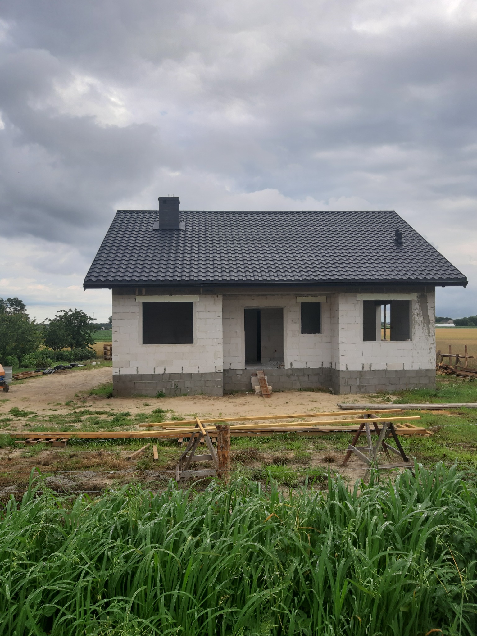 Surowy stan domu jednorodzinnego z białych bloczków, z czarnym dachem i kominem, widoczne otwory okienne i drzwiowe, na pierwszym planie trawa i drewniane elementy konstrukcyjne.