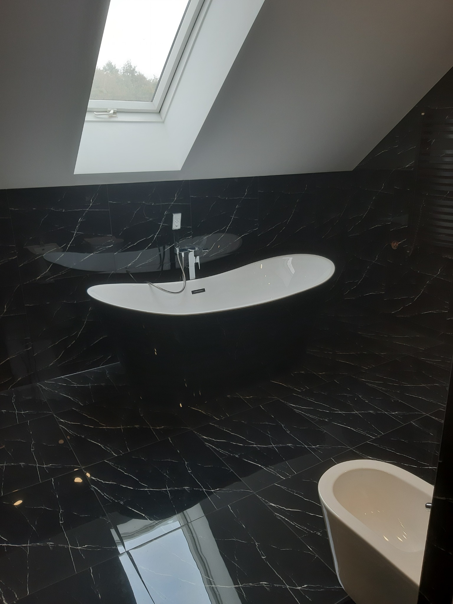 Nowoczesna łazienka na poddaszu z czarnymi marmurowymi płytkami, wolnostojącą białą wanną i oknem dachowym.