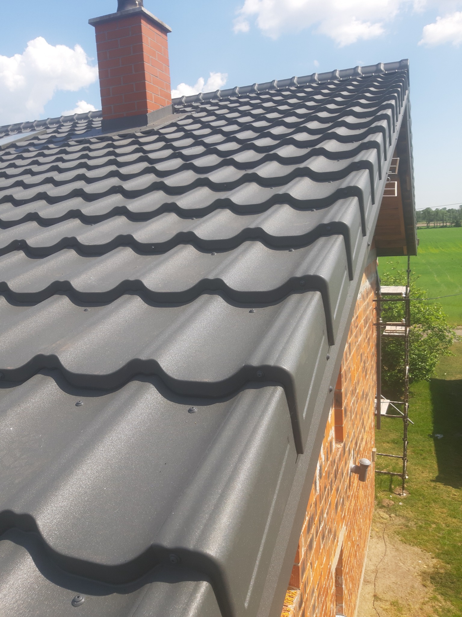 Szara blachodachówka na dachu murowanego domu z cegły, widoczny komin z cegły i rusztowanie przy ścianie.