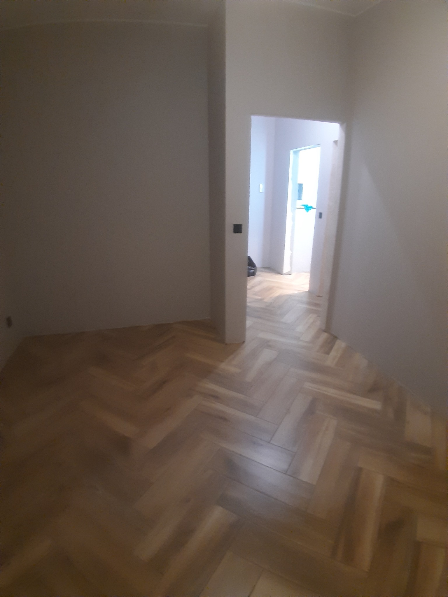 Wnętrze pokoju z parkietem ułożonym w jodełkę, widok na otwarte drzwi do kolejnego pomieszczenia, ściany w odcieniach bieli i beżu.