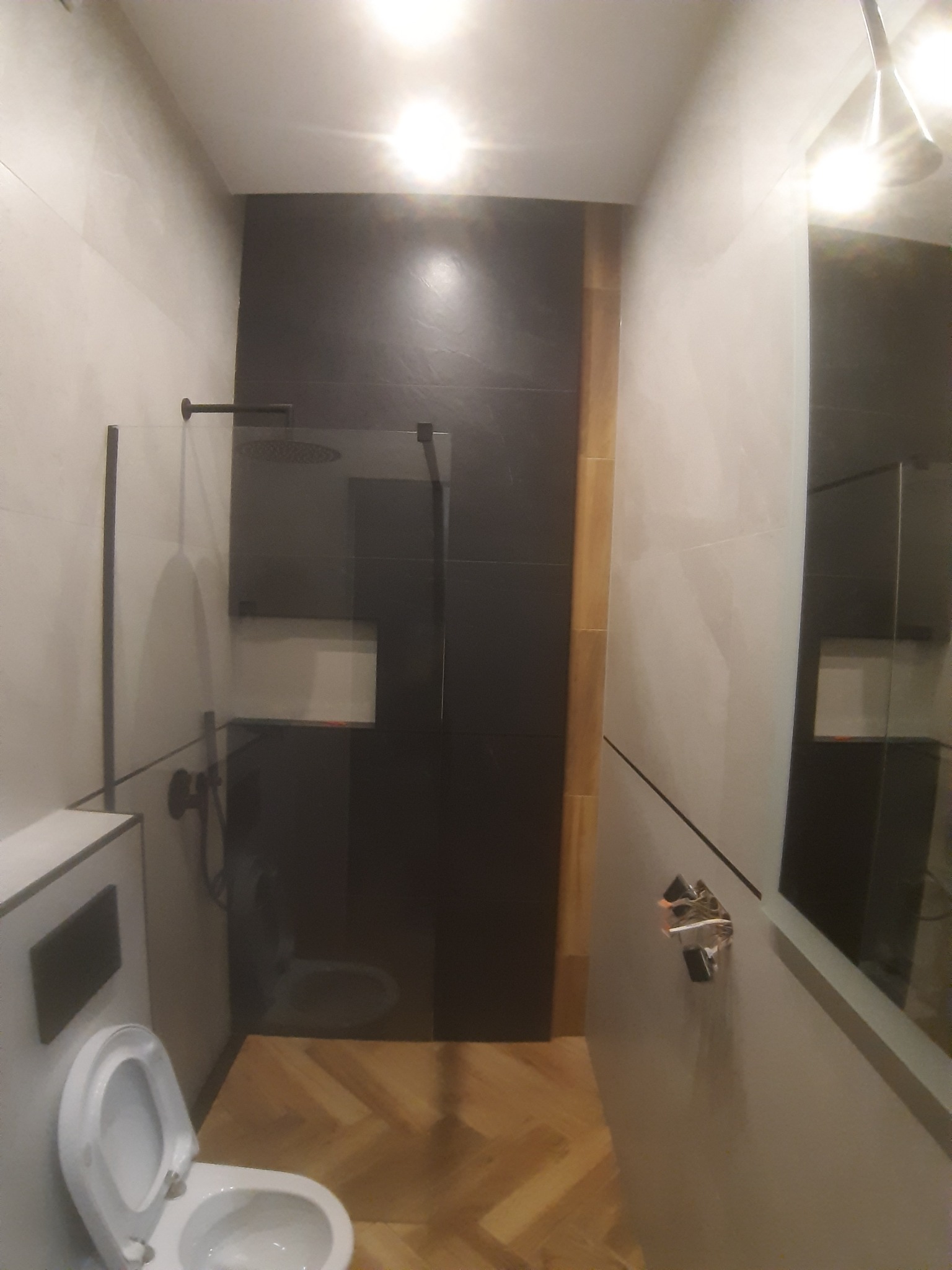 Nowoczesna łazienka z czarną kabiną prysznicową typu walk-in, białą ceramiką i podłogą z parkietu ułożonego w jodełkę, oświetlona jasnym światłem.