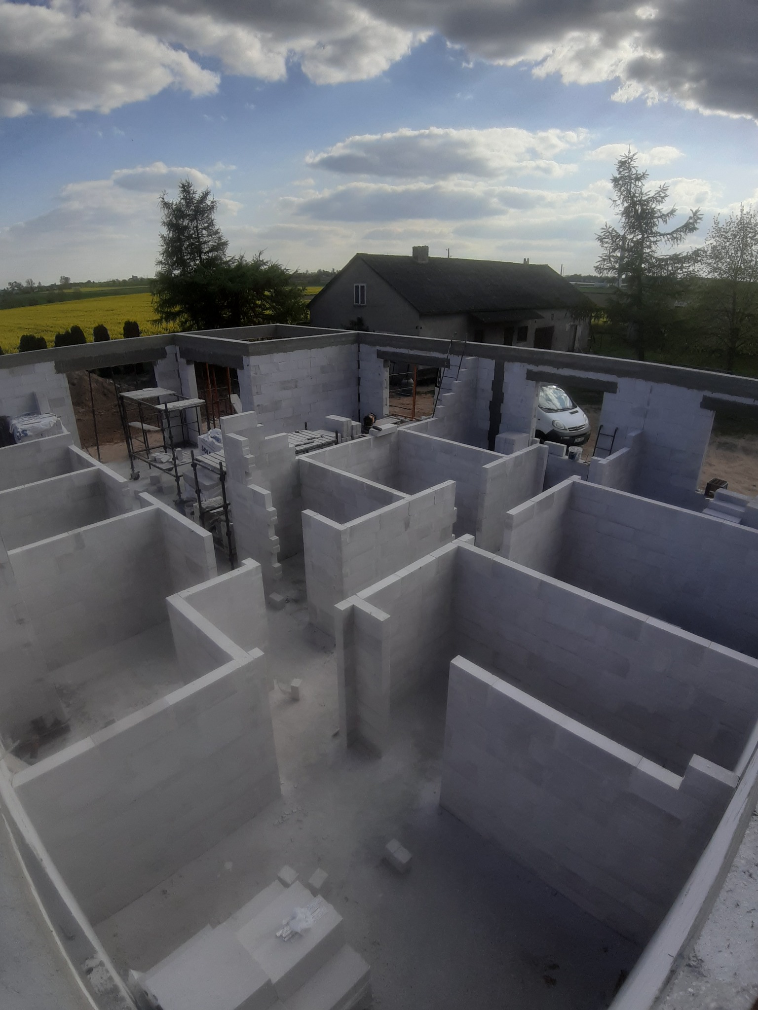 Wewnętrzne ściany domu w trakcie budowy wzniesione z białych bloczków, widoczne rusztowania i samochód w tle, żółte pole rzepaku i dom w oddali.