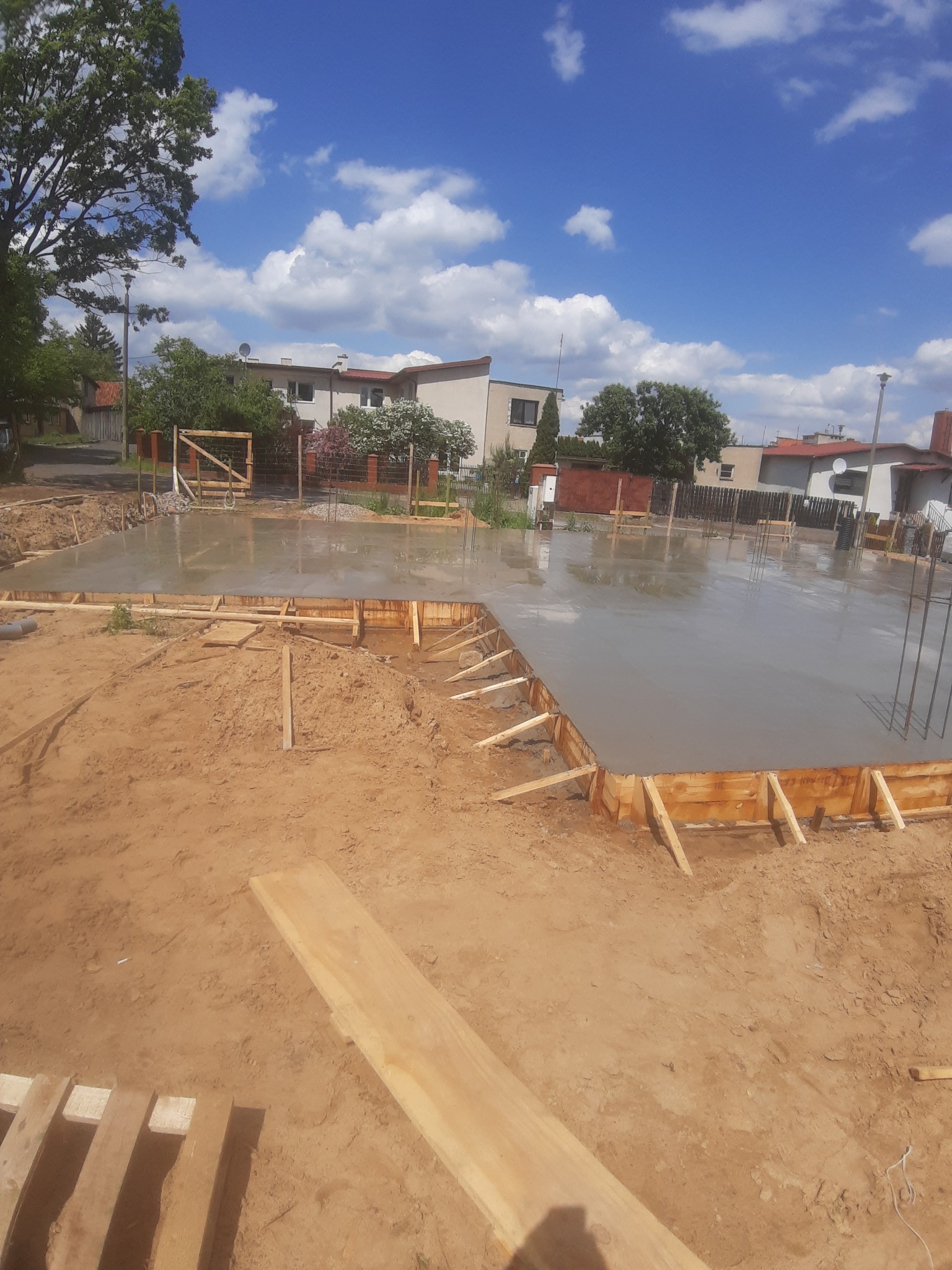 Wylany fundament z drewnianym szalunkiem w kształcie prostokąta na tle domów jednorodzinnych i błękitnego nieba.