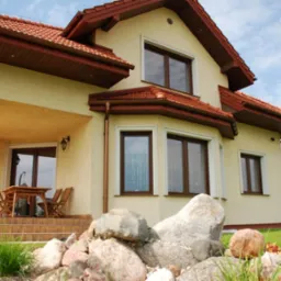 Elewacja domu jednorodzinnego z jasnego tynku, ciemnobrązową stolarką okienną i czerwonym dachem, widok z ogrodu z dekoracyjnymi kamieniami.