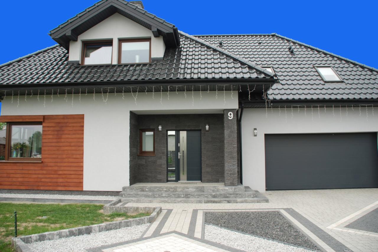 Nowoczesny dom jednorodzinny z szarą stolarką okienną i drzwiową, elewacją w odcieniach bieli i szarości, oraz numerem '9' nad wejściem, otoczony zadbanym trawnikiem i podjazdem z kostki brukowej.