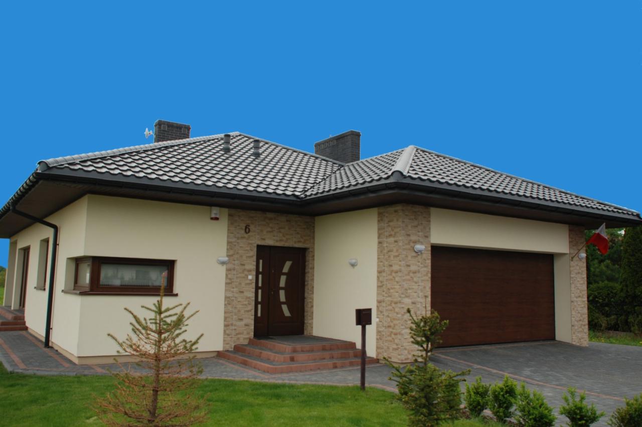 Nowoczesny parterowy dom z ciemnobrązowymi drzwiami wejściowymi i bramą garażową, jasną elewacją częściowo wykończoną kamieniem dekoracyjnym, szarą dachówką i zadbanym trawnikiem.