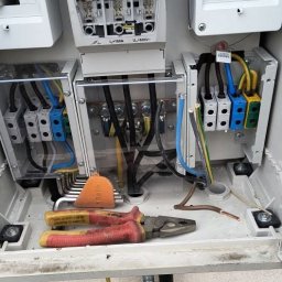 High Quality - Otwarta skrzynka rozdzielcza z bezpiecznikami APATOR 160A i wyłącznikami 25A, widoczne kolorowe przewody, narzędzia i uziemienie na białej ścianie.