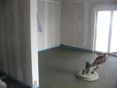 Szlifowanie świeżej, szarej posadzki w pomieszczeniu z przygotowanymi ścianami; widoczna maszyna szlifierska, taśma ochronna przy ścianach i okno balkonowe.