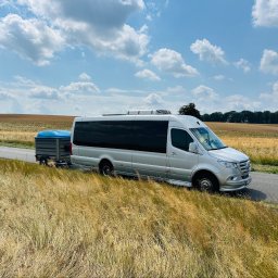 BUSEM- przewóz osób - Srebrny bus pasażerski z przyciemnianymi szybami i klimatyzacją dachową, ciągnący szarą przyczepkę, stoi na poboczu drogi wśród złotych pól pod błękitnym niebem.