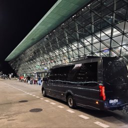 BUSEM- przewóz osób - Elegancki, ciemny bus Mercedes Sprinter z przyczepą, oczekujący na pasażerów z bagażami przed szklanym terminalem lotniczym w Gliwicach, oświetlony nocą.