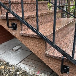 Granlux - Zewnętrzne schody z czerwonobrązowego granitu o nakrapianej fakturze, z czarną, kutą balustradą. Mokre stopnie prowadzą w górę, a u podstawy leżą dekoracyjne kamienie.