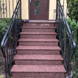 Granlux - Schody wejściowe z nakrapianego czerwono-różowego granitu, z czarnymi kutymi balustradami, prowadzące do brązowych drzwi z trzema przeszkleniami, na tle jasnożółtej elewacji.