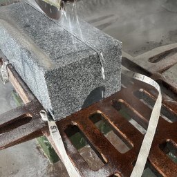 Granlux - Przemysłowe cięcie dużego bloku nakrapianego szarego kamienia piłą tarczową z intensywnym chłodzeniem wodnym, zamocowanego na zardzewiałej metalowej kratownicy białymi pasami.