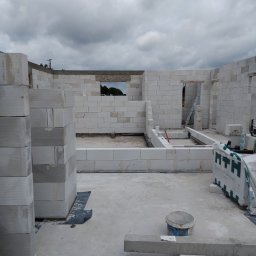 Patryk jankowski - Budowa ścian parteru domu z białych bloczków betonu komórkowego, widoczne otwory okienne i drzwiowe, na placu budowy wiadro i drabina, pod pochmurnym niebem.