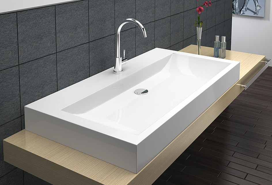 Biała prostokątna umywalka nablatowa z chromowaną baterią, zamontowana na drewnopodobnym blacie, na tle ciemnych, kwadratowych płytek ściennych i ciemnej podłogi.