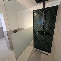 AB Construction - Nowoczesna kabina prysznicowa z ciemnozielonymi, błyszczącymi płytkami ułożonymi w jodełkę, czarną deszczownicą i częściowo matową szklaną ścianką. Jasne płytki na podłodze.