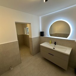 AB Construction - Nowoczesna łazienka z okrągłym lustrem LED, wiszącą szafką z umywalką i czarną armaturą. Ściany wykończone beżowymi płytkami wielkoformatowymi i jasną farbą.
